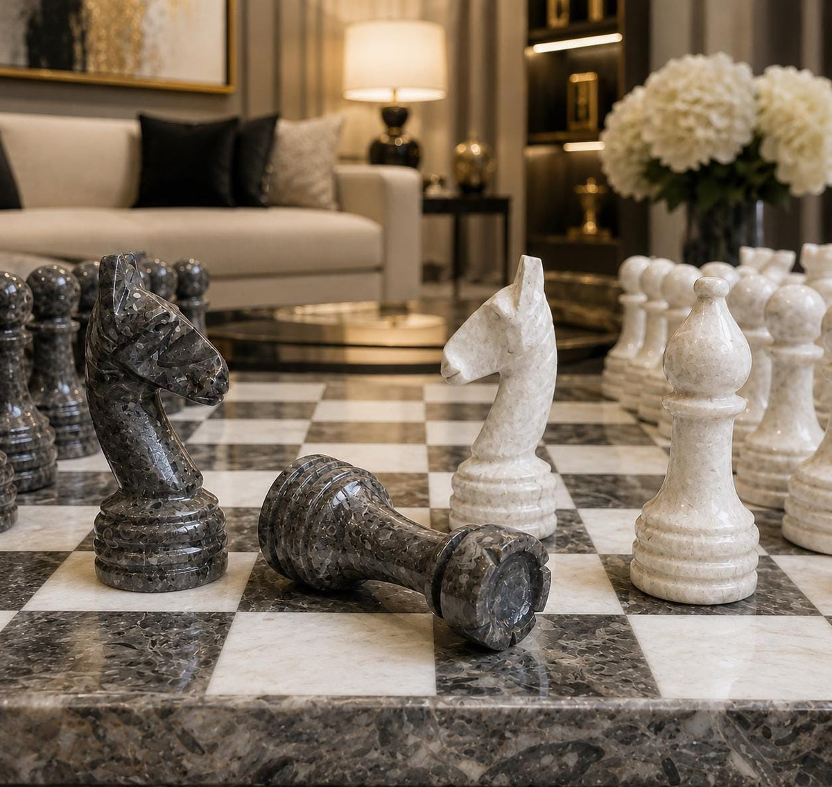 Large marble chess set on a checkered chessboard in a luxurious modern living room setting. The set features polished dark gray and off-white stone pieces, with one dark gray knight lying on its side and several off-white pawns lined up in the background.