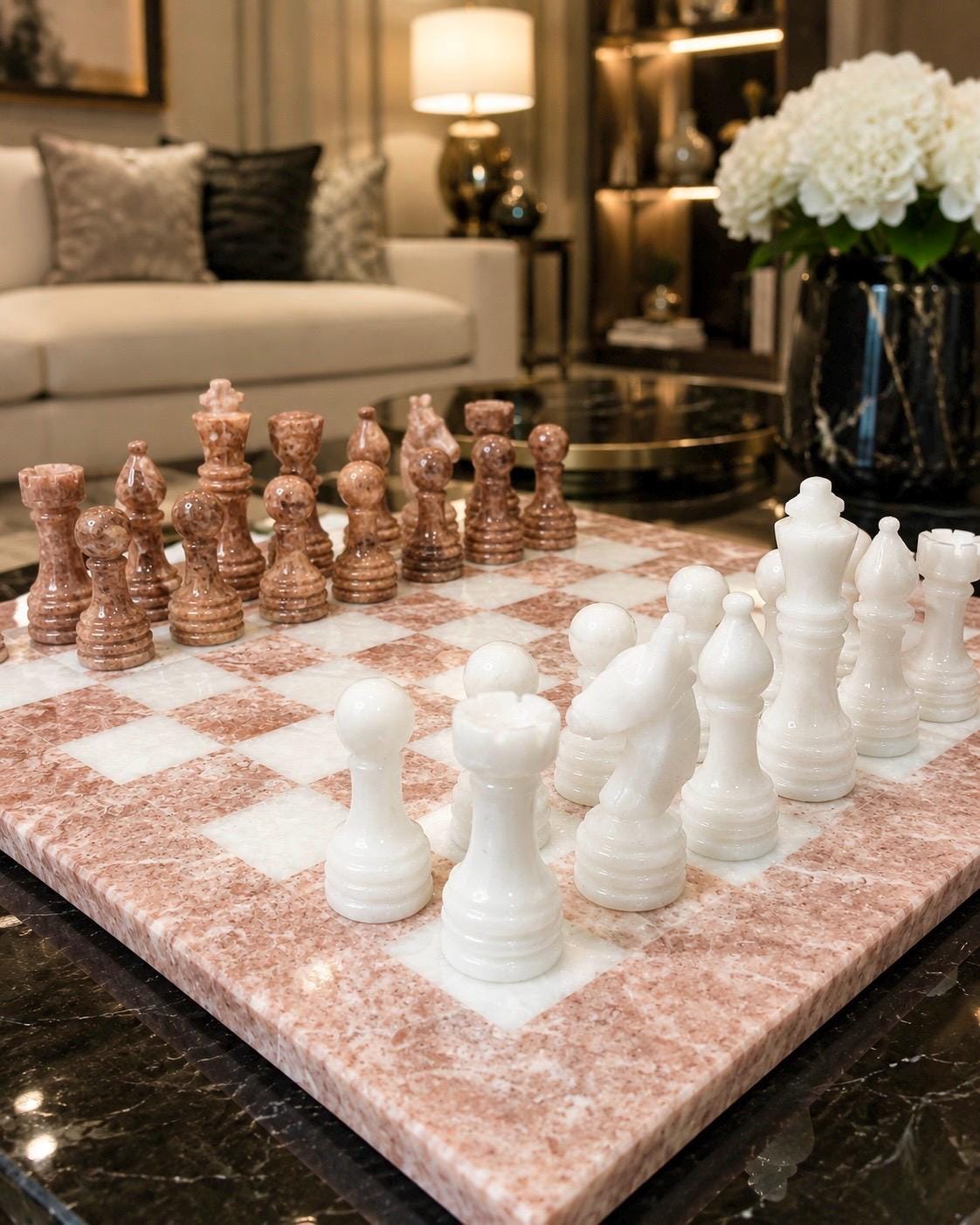 A marble chess set with white and pinkish-beige pieces on a matching marble board, placed on a dark marble table in a modern living room setting.
